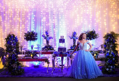 Debutante de vestido azul diante da decoração da festa de 15 anos, com flores azuis e cortina de luzes