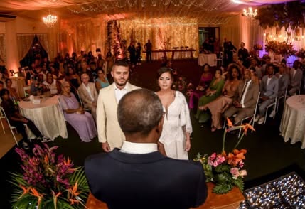 Cerimônia de casamento vista do altar: noivos diante do celebrante, com convidados e salão iluminado por lustres